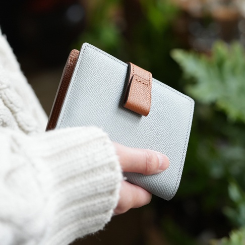 Atelier nuu noble 皮革雙折短夾，淺灰色荔枝紋皮革搭配棕色繫繩，由手持於柔和的自然光線下。