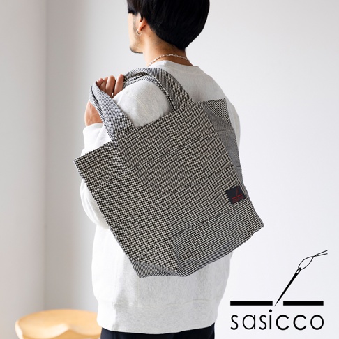 一款採用三河木綿刺子織材質的黑色與白色格紋大容量托特包，由一位男性從側面背起，包包上有一個紅色小標籤。