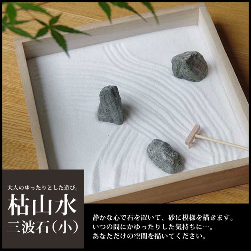 日式枯山水模型，木製淺盤內盛白色細砂，砂上耙出波浪紋，擺放三顆灰色不規則岩石，其中一顆旁有一迷你木耙。