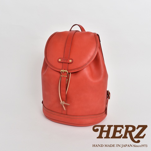 橘紅色皮革單扣束口後背包，設有寬版提把、束繩開口、品牌標誌、金屬扣環和車縫線裝飾。