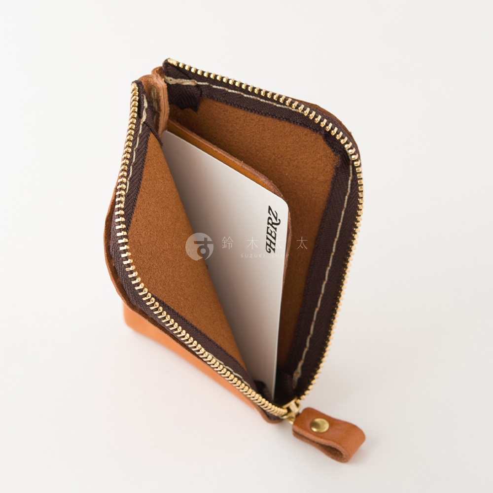 這款由日本HERZ職人手作的咖啡色皮革迷你零錢包，採L型拉鍊設計，內部設有夾層並放入了一張印有HERZ品牌字樣的白色卡片，整體呈現簡約精緻的質感。