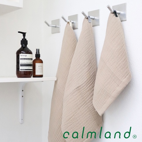 三條米色無撚紗質長方巾掛在牆上的掛鉤上，左側架子上擺放著兩瓶棕色瓶裝的洗浴用品。
