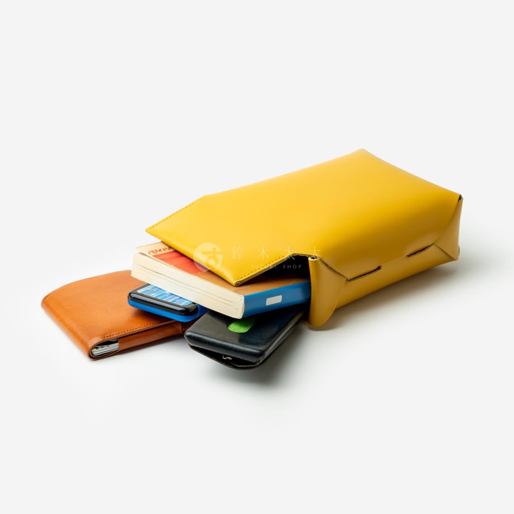 FABRIK 經典皮革可拆式多用途迷你斜背包，黃色包款側躺，內含棕色皮夾、兩支手機和一本藍色封面的書籍，背景為純白。