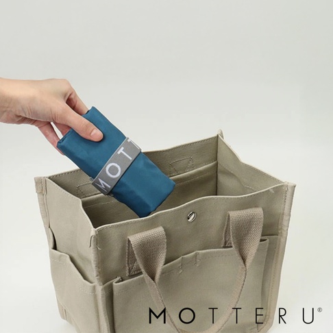 一隻手將一個藍色摺疊保冷袋放入一個卡其色帆布托特包內。摺疊袋上繫著一個灰色束帶，上面印有白色「MOT」字樣。