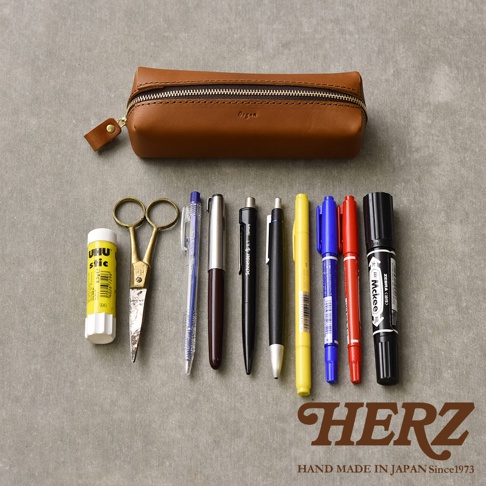 職人手作的棕色皮革Organ箱型收納筆袋，搭配銀色拉鍊，袋上印有「Organ」字樣。筆袋前方擺放著一管黃色UHU膠水、一把金色剪刀，以及一系列不同顏色和款式的筆，包括藍色、銀色、黑色、黃色、藍色、紅色和一支黑色簽字筆。