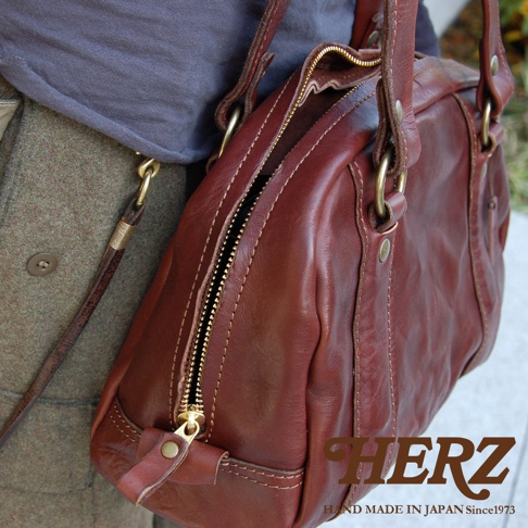 深棕色水洗皮革迷你波士頓包，設有金色拉鍊和金屬配件，搭配灰色上衣與橄欖綠外套。