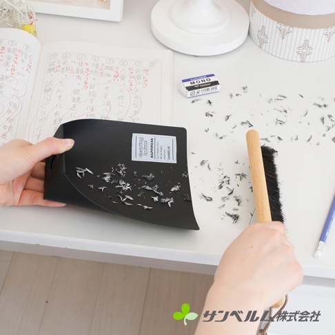 一張照片顯示一雙手正在使用木柄黑毛掃把與黑色畚箕清理桌面的橡皮擦屑。桌面上放有一本打開的日文練習簿和一個 MONO 橡皮擦，畚箕上印有英文標語，畫面右下角帶有サンベルム株式会社的品牌標示。