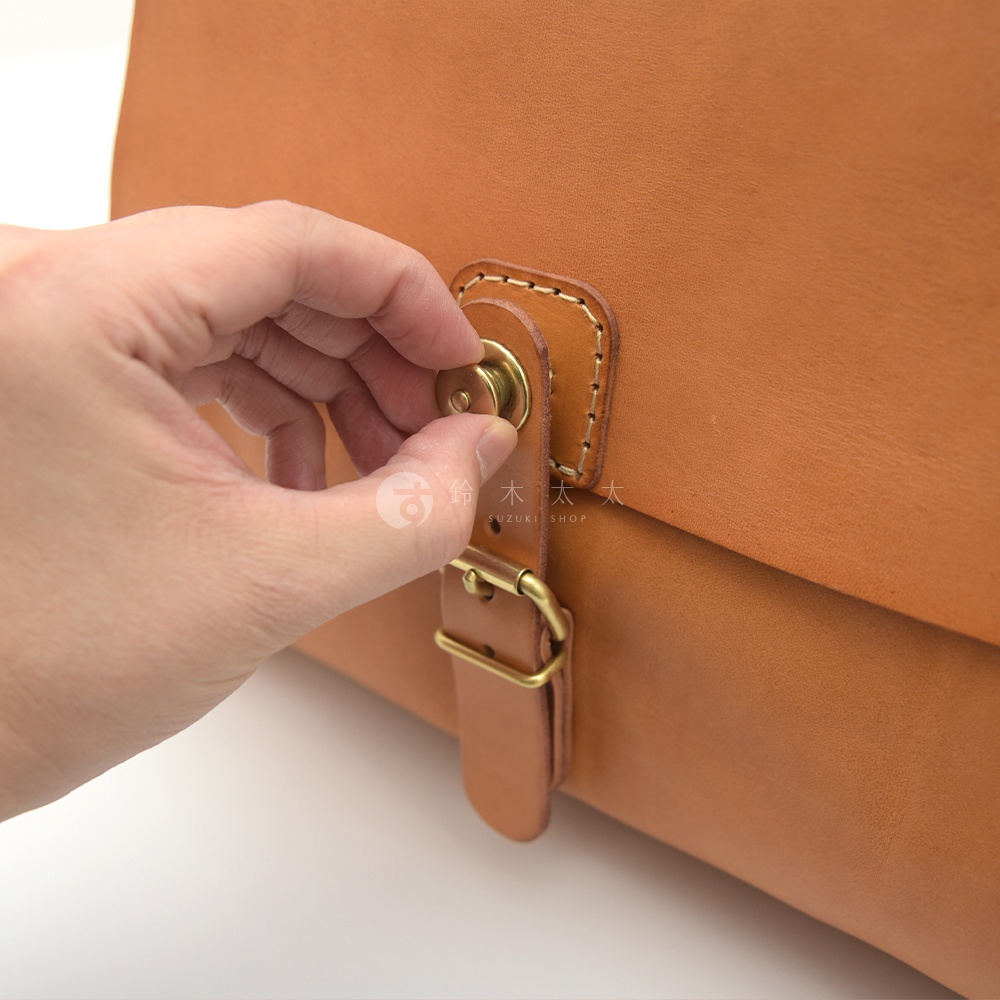 手持淺棕色牛皮後背包的旋鈕鎖扣，展示其精緻縫線和黃銅金屬件，皮革材質呈現自然紋理。