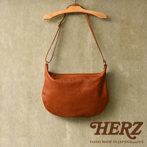 職人手作的駝色皮革單肩半月包，以自然垂墜的姿態掛在木質衣架上，包體飽滿，展現皮革的細緻紋理與光澤。