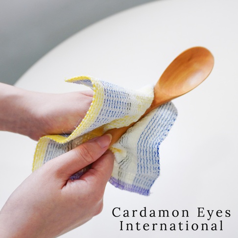 雙手拿著一條壓克力纖維無痕清潔巾，清潔巾為白色基底，織有藍色細條紋和黃色邊緣，正在擦拭木湯匙。