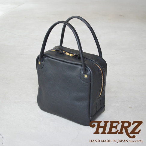 一個由日本品牌HERZ手工製作的黑色皮革方形托特包，包身線條簡約俐落，配有圓形皮革提把與金色的金屬拉鍊，右下角印有HERZ品牌標誌與創業年份。