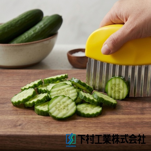 一隻手握著黃色握柄的下村工業波浪造型切刀，正在木質砧板上切開小黃瓜，砧板上已有許多切好的波浪狀黃瓜片，背景則有一個裝著整根黃瓜的碗與一個小碟子。