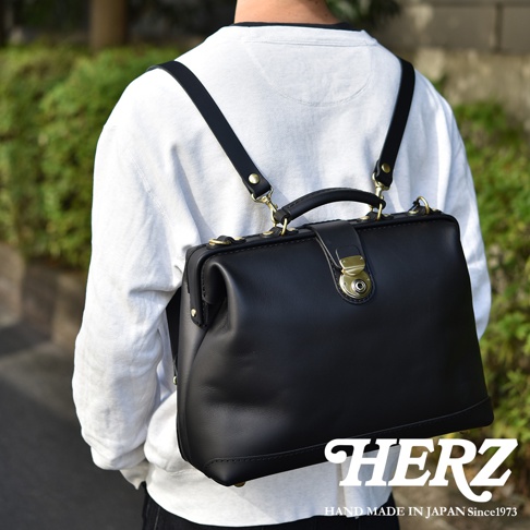 一位穿著白色上衣的模特兒，背著一個日本HERZ手工製作的黑色皮革杜勒斯公事包，包款採用經典口金設計搭配復古黃銅五金扣環，背帶可拆卸轉換為背包或手提模式。