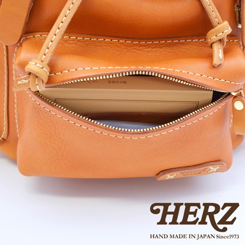 職人手作皮革復古風旅行後背包 R-65，特寫展示經典棕色皮革材質，帶有精緻的白色縫線。拉鍊部分敞開，露出內袋中的書本與卡夾。