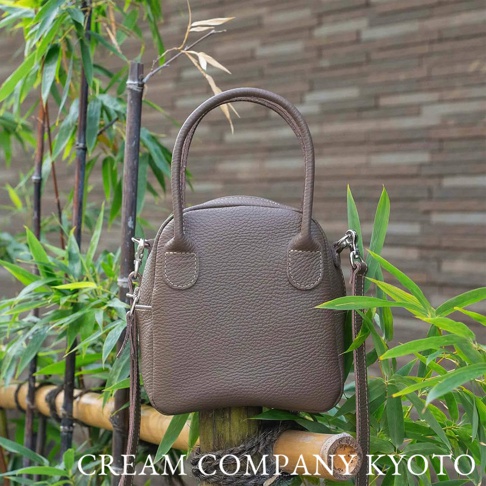 深棕色皮革手提包，貝殼造型，有頂部提把和可拆卸的斜背包帶，包身有縫線裝飾，置於竹子和綠葉背景前。