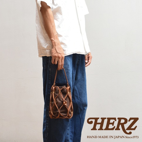 一位男子穿著白色襯衫和牛仔褲，手裡提著一個棕色編織皮革水桶包，背景為白色牆壁，下方有「HERZ HAND MADE IN JAPAN Since1973」的文字。