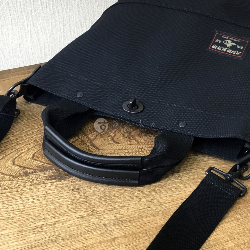 黑色帆布兩用托特包，配有黑色皮革手柄和可拆卸肩帶，包身設有金屬旋扣和品牌標籤。