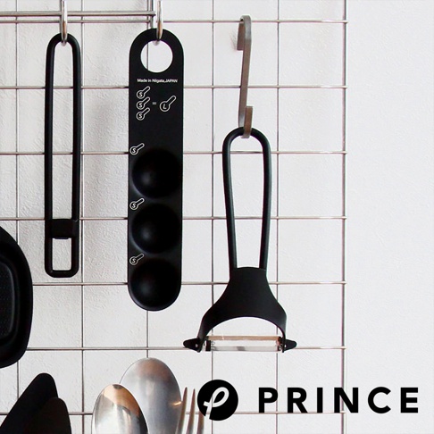 黑色削皮器，設有彎曲的黑色手柄和銀色刀片，掛在金屬網架上。