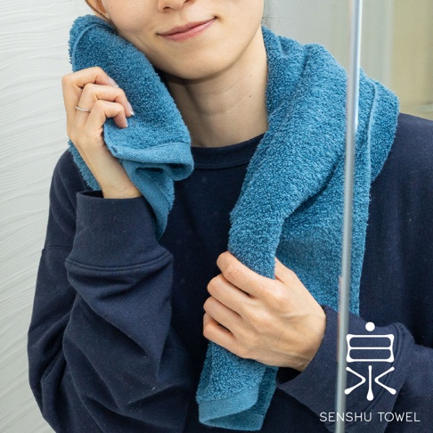 一名女子將藍色長纖毛巾搭在肩上，一端輕柔地擦拭臉頰。毛巾質地蓬鬆，呈現溫柔的藍色調。