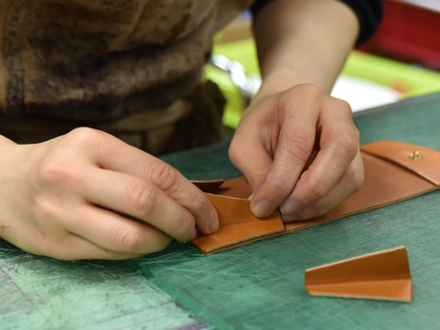 職人正在手作一個棕褐色皮革雙折收納零錢包，將一片皮革對折，另一片皮革準備縫合。