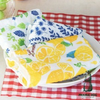 三條五層紗蚊帳質地擦拭巾，分別印有綠色植物、藍色花紋及黃色檸檬圖案，質地柔軟吸水，疊放在白色盤子裡，背景為紅色格紋桌布。