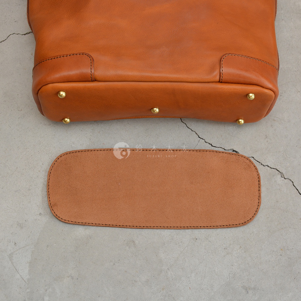 以駝色皮革製成的日本HERZ Organ兩用拉鍊托特包局部特寫，包包底部裝有黃銅腳釘，下方擺放著同色系的皮革襯墊