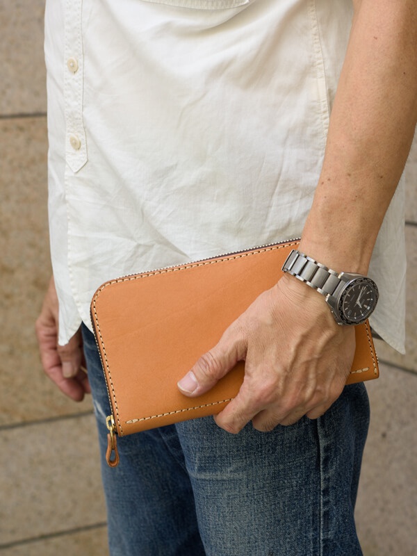 淺棕色皮革拉鍊長夾，以米白色縫線勾勒邊緣，搭配金色拉鍊頭。由男性手持，搭配淺藍色牛仔褲與白色襯衫。