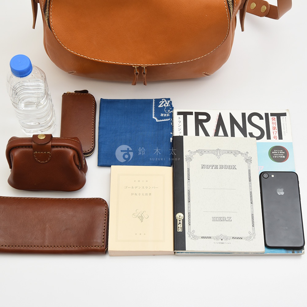 HERZ職人手作皮革大容量馬鞍包 CK-150，搭配水瓶、皮夾、筆記本、手機和一本名為「ゴールデン スランバー」的書，整體呈現在白色背景上。