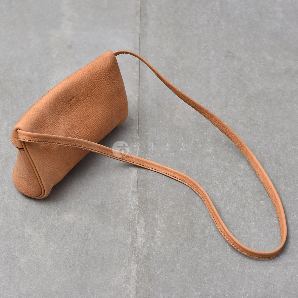 一個淺棕色皮革製成的貼身斜背包，包身呈現圓潤的長條狀，搭配細長的同色系皮革背帶，包包上印有品牌標誌，整體擺放在灰色水泥紋理的背景上。