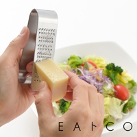 雙手持用不鏽鋼立式磨泥器，將一塊淺黃色起司磨碎，背景為一碗色彩繽紛的沙拉。