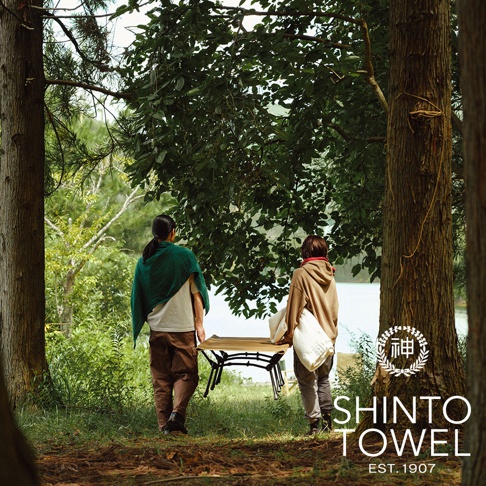 兩名人士在林間湖畔，其中一人肩披綠色織品，另一人攜帶白色毛毯，旁邊有一張可折疊的桌子。