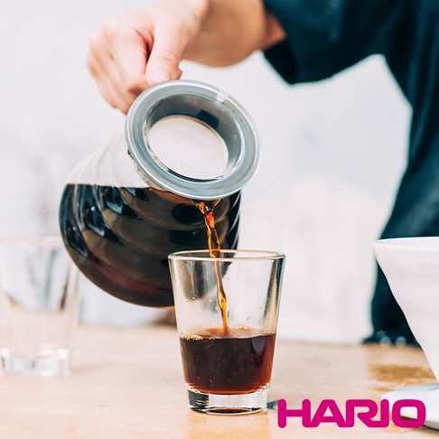 一個玻璃咖啡壺正在將深棕色咖啡倒入一個透明玻璃杯中，咖啡壺帶有銀色金屬濾網和透明瓶蓋。