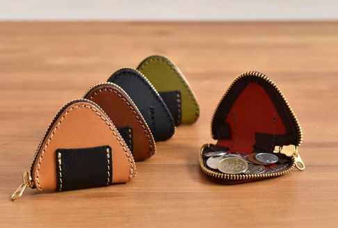 職人手作皮革三角飯糰造型零錢包，有淺棕色、棕色、黑色、綠色等顏色。淺棕色零錢包的正面有黑色皮革裝飾，並以白色縫線點綴。其中一個零錢包打開，展示其紅色的內襯和裝有硬幣的樣子。