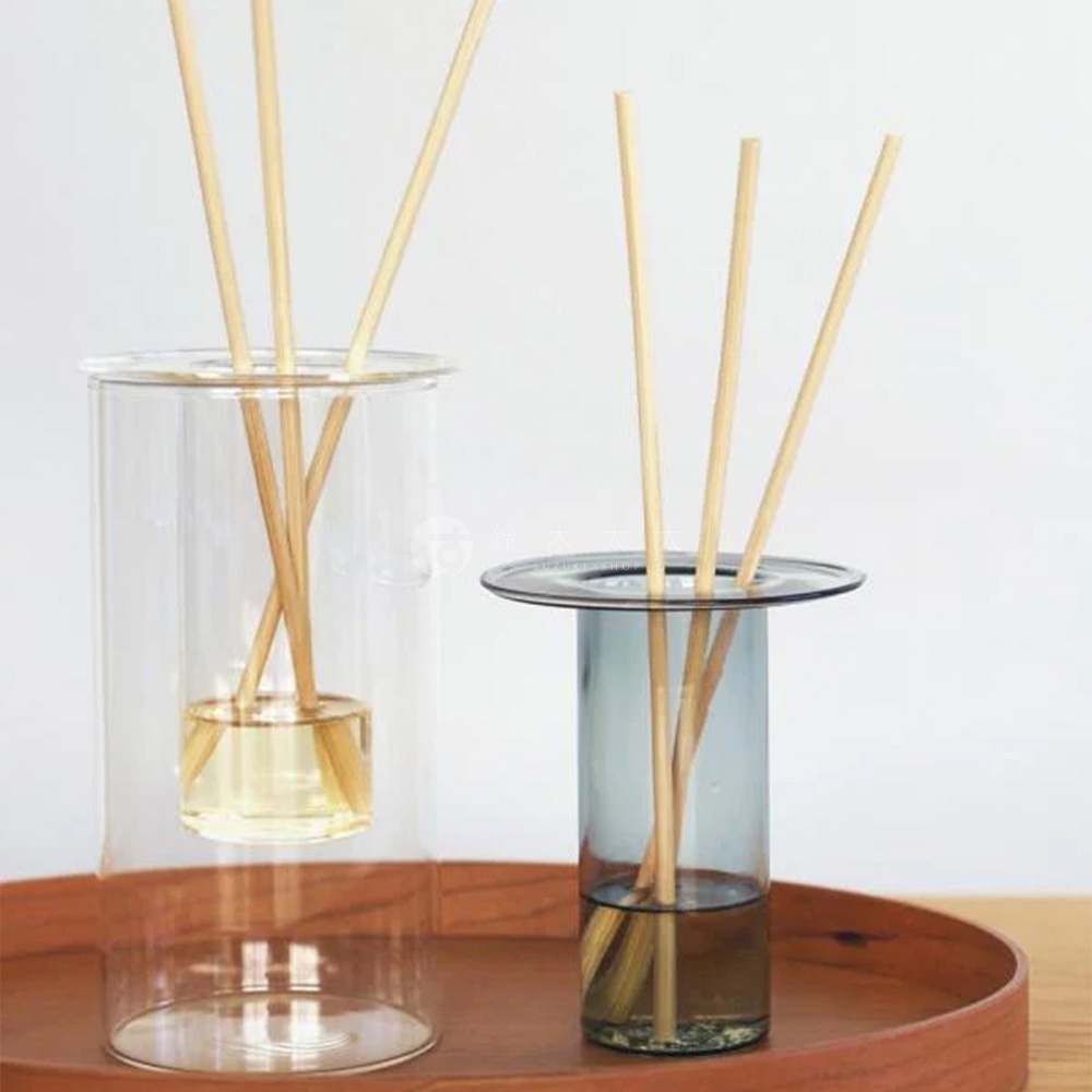 玻璃花瓶和擴香棒，左邊的花瓶是透明玻璃，裝有黃色液體，右邊的花瓶是灰色玻璃，裝有深色液體，擺放在木質托盤上。