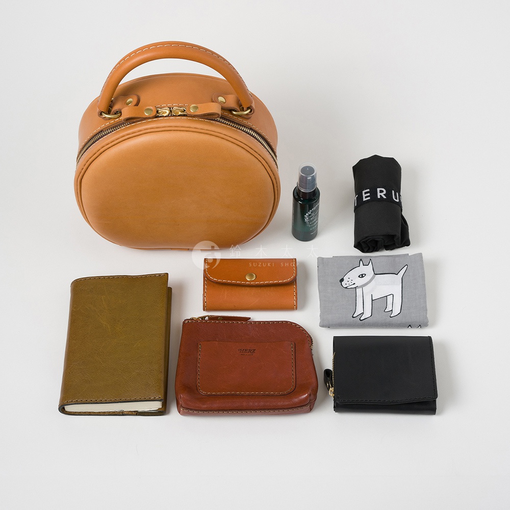 一個日本製HERZ品牌皮革材質的圓形手提包，旁邊擺放著一個棕色零錢包、一個黑色錢包、一個筆記本、一個小型噴霧瓶和一個印有白色小狗圖案的淺灰色布料。