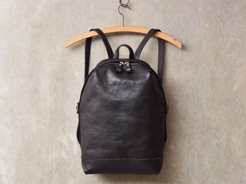 深棕色皮革後背包，簡約褶皺設計，配有雙拉鍊主袋，皮革材質，掛於木質衣架上，背景為淺色紋理牆面。