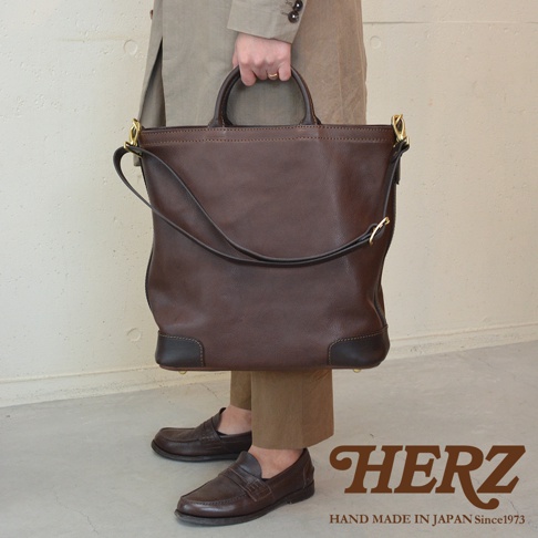模式B： 日本HERZ職人手作皮革Organ兩用拉鍊托特包，深棕色，雙提把和可拆卸斜背帶，搭配棕色樂福鞋，背景牆為米白色，地面為灰色。