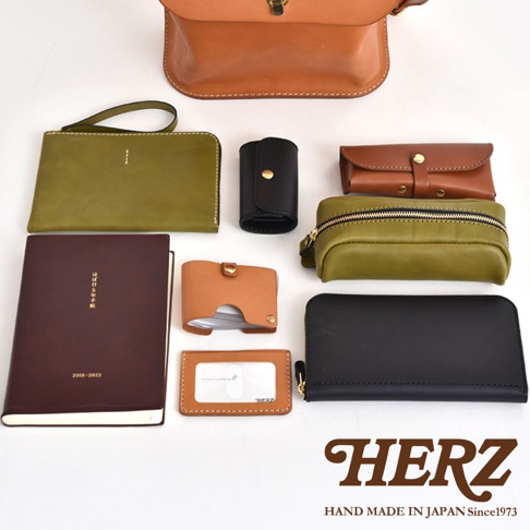 展示多款職人手作皮革配件，包含棕色翻蓋包、綠色手拿包、黑色鑰匙圈、棕色零錢包、綠色筆袋、深棕色筆記本、兩款卡夾及黑色長夾。