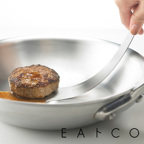 日製金屬食物鏟，鏟面上放著一個煎好的漢堡肉排，肉排表面有醬汁，鏟子伸入鍋中，鍋身為不鏽鋼材質。