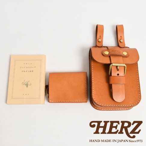 棕色皮革腰袋，帶有黃銅扣環和皮帶。旁邊有一個小巧的棕色皮革錢包和一本淺黃色封面書籍，書名為「ジャイロスコープ」。