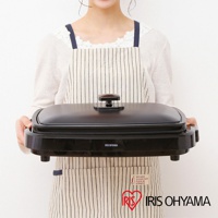 黑色烤網風電烤盤 APA-136，帶有方形黑色提手，置於條紋圍裙前，品牌標誌「IRIS OHYAMA」於產品正面中央。