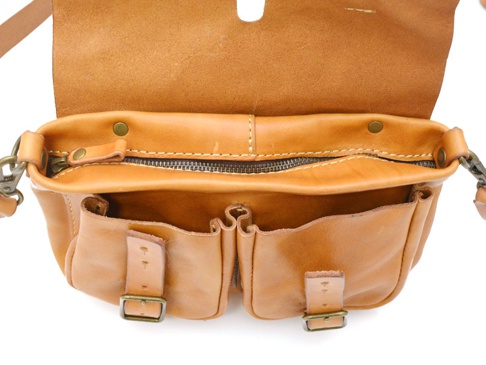 淺棕色皮革肩背包，具有兩個外部口袋，每個口袋都有皮帶和金屬扣環。包包上方有拉鍊開合，內裡為皮革材質。