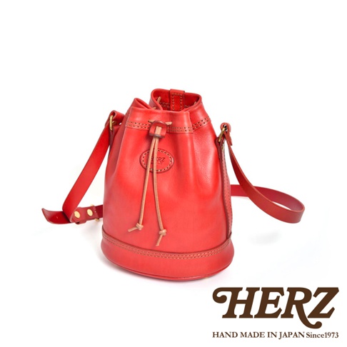 紅色職人手作皮革束口單肩包，圓筒狀包身，附有可調整的皮革背帶和束繩開口，包身底部有縫線裝飾，品牌標誌壓印在包身正面。