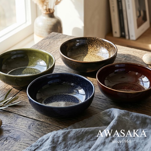 放置在木質桌面上的四個AWASAKA品牌多用途寬口湯碗，分別呈現出綠色、褐色、深藍色與赤褐色釉面，碗身帶有細緻的斑點紋理與光澤感，右下方擺放著灰色亞麻布，畫面整體風格簡約雅緻。