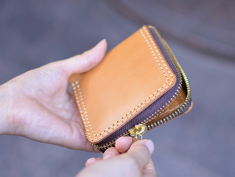 一隻手拿著一個棕褐色皮革製作的短夾，拉鍊部分有深棕色布邊與金色拉鍊頭，縫線為白色。