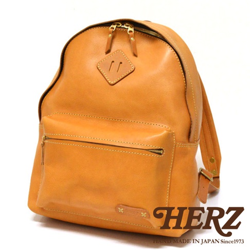 職人手作皮革休閒風後背包，採用鞣製皮革製成，呈現自然棕色調。包款正面設有拉鍊式收納袋，頂部有雙拉鍊設計，並點綴菱形皮革飾片。整體風格簡約，適合日常休閒穿著。
