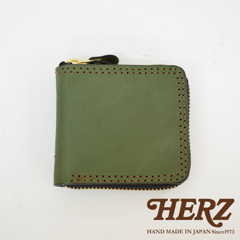 正面視角展示一個日本HERZ品牌製作的橄欖綠色手工皮革短夾，皮夾外圍以深色縫線裝飾並配有金色拉鍊，整體設計簡約厚實，右下角印有品牌標誌與創立年份。