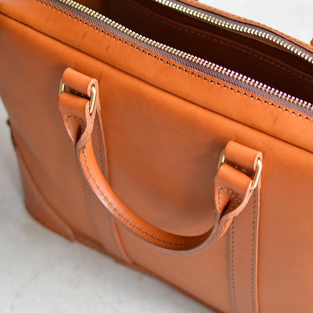 職人手作皮革Organ商務公事包 G-40，特寫鏡頭展示了光滑的淺棕色皮革、縫線細節，以及金屬拉鍊和提把上的黃銅扣環。
