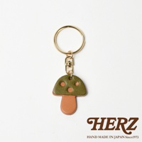 職人手作皮革香菇造型鑰匙圈，傘蓋為綠色皮革，帶有三個橘色圓點裝飾，傘柄為棕色皮革。搭配金色鑰匙圈和掛鉤，整體呈現可愛的蘑菇造型。