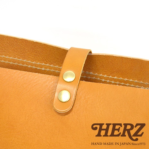 日本HERZ職人手作皮革軟皮橫式斜背包，特寫皮革紋理、縫線細節與黃銅色釦具，並展示品牌Logo。